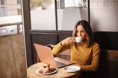 Kafedeki dizüstü bilgisayarda çalışırken kahve içip kruvasan yiyen genç bir kadın, Kafkasyalı bir kadın internetteki uzaktan kumandayla kahvaltının tadını çıkarıyor.