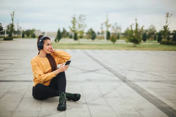 スマートフォンで音楽を聴くヘッドフォンの幸せな若い女性の屋外写真と笑顔空のスタジアムで道路に座っている間 カジュアルな服を着て — ストック写真