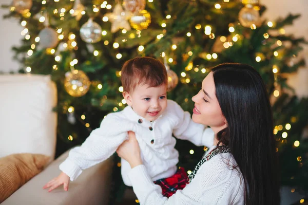 若いです女性笑顔と遊びます可愛いです男の子とともにぼやけた背景の装飾された針葉樹のクリスマスの日に自宅で — ストック写真
