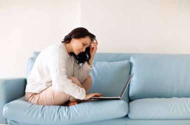 Sıradan giyinmiş genç bir kadın dizüstü bilgisayarında klavyesiyle kanepeye oturmuş, bilgisayar ekranına bakıyor, önemli bilgiler arıyor ya da acil e-postaları cevaplıyor.