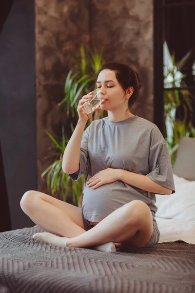 Hydration during pregnancy. Young pregnanat woman in t-shirt sitting on bed at home, drinking clean mineral water in morning to prevent dehydration. expectant mother staying healthy and hydrated
