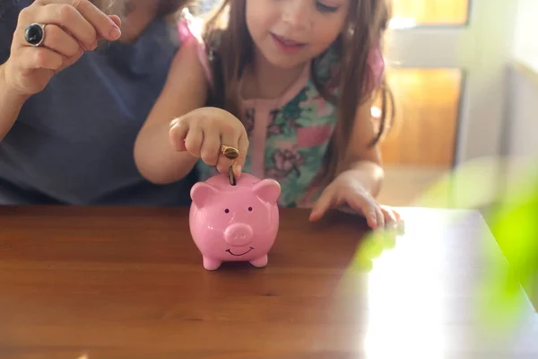 小さな女の子は家で母親とテーブルに座っている間に貯金箱にコインを入れて 女性は子供のための予算の基本を説明し 賢明にお金を予算する方法を子供に教えています 金融教育 — ストック写真