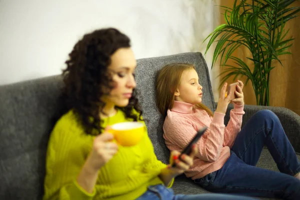自宅のソファで別々に座っている間 携帯電話を使用している若い母親と女の子の娘に焦点を当て 手元のガジェットとソファでリラックスした子供に焦点を当てます 家庭生活における技術 — ストック写真