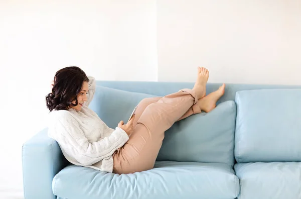 年轻的黑发女子躺在舒适的沙发上 姿势放松 双腿翘起 手持手机 凝视着屏幕 在平静的家庭环境中在线聊天 阅读重要的信息 图库图片