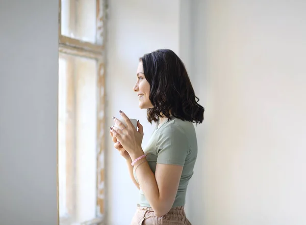 西德威的照片上 年轻快乐的黑发女子站在室内喝茶 看着窗外 温柔地微笑着 看着外面的天气 是时候放松一下 享受一下家的舒适了 — 图库照片