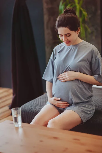 怀孕期间的健康习惯 年轻快乐的孕妇在家里 一边坐在餐桌旁 一边喝着一杯清澈的水 一边轻柔地抚摸着肚子 喝着水来维持身体和婴儿的健康 — 图库照片