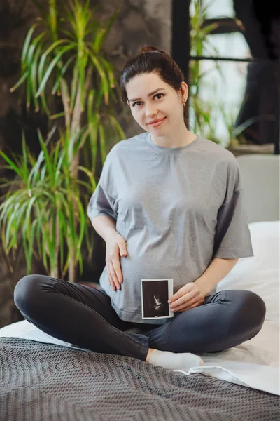 妊娠超音波を保持しながら 妊娠中の女性は 自宅でベッドの上にハスのポーズに座って 赤ちゃんソノグラムを持つ妊婦 産後ケア — ストック写真