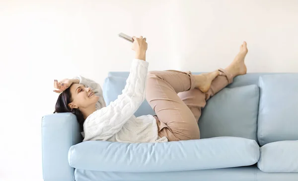 年轻的黑发女子躺在舒适的沙发上 姿势放松 双腿翘起 手持手机 凝视着屏幕 在平静的家庭环境中在线聊天 阅读重要的信息 免版税图库图片