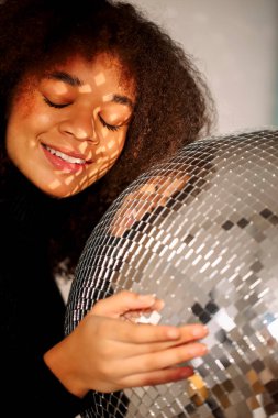 Genç, mutlu, Afro-Amerikan kıvırcık kızı gözlerini zevkle kapatırken elinde kocaman gümüş bir disko topu, gülümseyen, kulüp atmosferinde parti dekorasyonu yapmış, gevşemiş siyah kıvırcık kız. Gece hayatı.