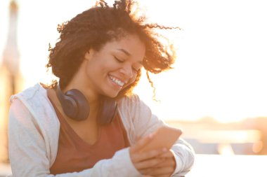 Açık havada, akıllı telefon kullanan ve mutlu bir şekilde gülümseyen mutlu Afro-Amerikan kadın portresi. Sabah güneşinde çatıda dikilen, cep telefonlu neşeli melez kadın.