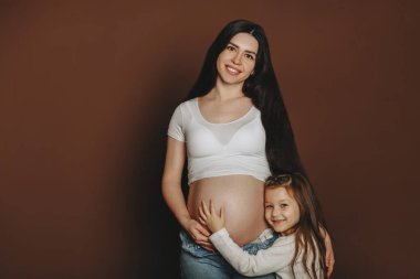 Genç, mutlu bir ailenin, hamile bir kadının ve küçük tatlı bir kızın stüdyo portresi. Annesinin karnına nazikçe sarılan bir kız çocuk bekliyor. Kardeşlerini bekliyor. Ebeveynlik ve annelik kavramı