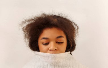 Bej arka planda izole edilmiş üzgün, üzgün, Afro-Amerikan kıvırcık kızının portresi. Üzüntülü siyah bir kadın, ağzı örgü süveterle kapatıyor ve aşağıya bakıyor. Yakından. Üzüntü ve yalnızlık