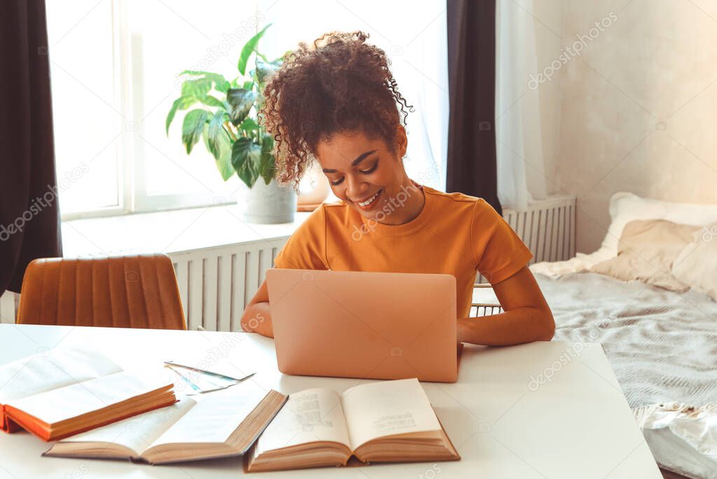 Cheerful young African American female student sitting at desk at home surrounded by textbooks, in front of laptop studying hard prepares for exams online, looking in laptop with satisfied smile