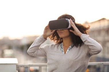 Geleceğin teknolojisi. Sanal gerçekliği keşfeden, arka planda şehir manzarası olan, sanal gerçekliği araştıran, sanal dünyayla etkileşime geçmeye çalışan, VR gözlüklü, modern melez genç bir kadın.