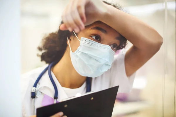 年轻疲惫的过度劳累的非洲裔美国医务工作者戴着防护面罩 手里拿着X光扫描 带着沮丧的表情望着窗外 Covid 19大流行病期间的保健工作者 — 图库照片
