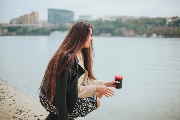 秋の日に川の水の近くにスクワットカジュアル服で手にコーヒーを取る若い女性が背景に街並みと 自然の中でレクリエーションを楽しんで海岸沿いのロマンチックな夢の女性 — ストック写真