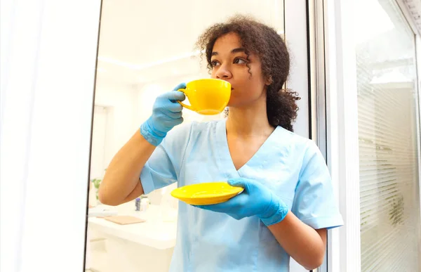 年轻的非洲裔美国女医生 身穿制服 戴着手套 一边喝咖啡或喝茶 一边在诊所看病 一边休息 在医院的艰苦工作日休息 — 图库照片