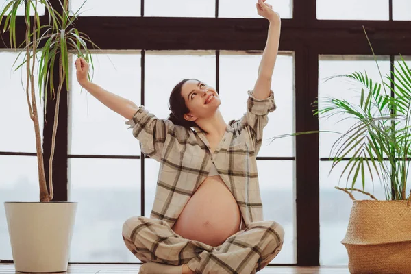 全身而退的年轻沉着的孕妇在睡梦中抱着肚子 坐着荷花 靠着全景窗 孕妇轻轻地摸着肚子 怀孕期间的沉思 — 图库照片