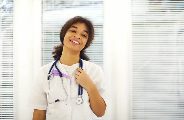 在现代诊所工作期间 身穿军服脖子上戴听诊器的快乐的非洲裔美国女医生对着摄像机微笑 图库图片