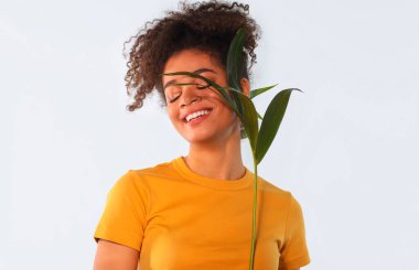 Genç mutlu Afro-Amerikalı kadın, kabarık saçlı, tropikal bitkileri yüzüne yakın tutan ve kapalı gözlerle gülümseyen, yeşil palmiye dallı hoş siyah kız, açık arkaplanda soyutlanmış.