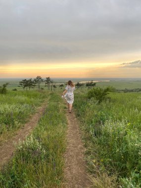 Yazın günbatımında yeşil çimlerin üzerinde koşan, uzun romantik elbiseli bir kadının arka planında, kaygısız, mutlu bir kadın çayırda yürüyor ve vahşi doğada eğleniyor.