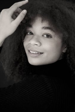 Çekici gülümseyen kıvırcık saçlı, siyah elbiseli, karanlık stüdyo arka planında yalnız poz veren Afro-Amerikan kadının portresi. Makyajsız, doğuştan güzel bir Afrikalı kadın model. Siyah ve beyaz