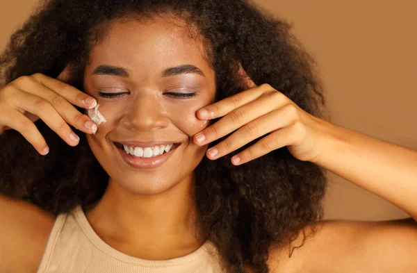 Smiling dark skinned woman with curly afro hair applies face cream on cheeks with both hands, poses over beige studio wall background, uses cosmetic product. Skin care and beauty treatments