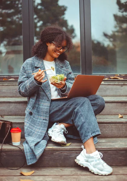 快乐的非洲裔美国女人吃着沙拉 一边吃着健康的午餐 一边在户外用笔记本电脑休息 秋天的一天 在网上学习的黑人女生在公园里休息 图库照片