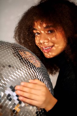 Genç, mutlu, Afro-Amerikan kıvırcık kızı gözlerini zevkle kapatırken elinde kocaman gümüş bir disko topu, gülümseyen, kulüp atmosferinde parti dekorasyonu yapmış, gevşemiş siyah kıvırcık kız. Gece hayatı.