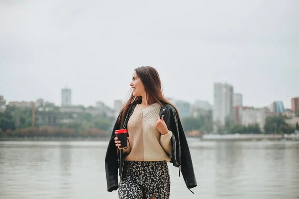 秋の日は川沿いの水で気軽にコーヒーを手に取る若い女性が街並を背景に ロマンチックな夢の女性が自然の中でレクリエーションを楽しむ海岸線 — ストック写真