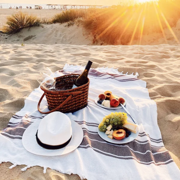 素晴らしい夕方の夕日の砂浜でロマンチックなピクニック ウィッカーバスケットのワインボトルとグラスで毛布 食べ物 果物やプレート上の軽食 — ストック写真