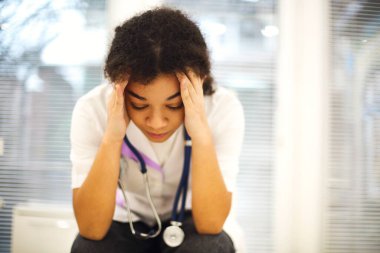 Üzgün, genç, afro-amerikan hemşire sinirli bir yüz ifadesiyle oturuyor, klinikteki iş yoğunluğundan bunalmış hissediyor, yorucu vardiyadan sonra yorgun hisseden etnik bayan doktor.