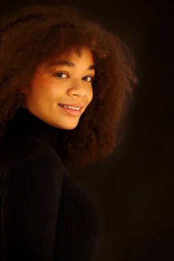 Çekici gülümseyen kıvırcık saçlı, siyah elbiseli, karanlık stüdyo arka planında yalnız poz veren Afro-Amerikan kadının portresi. Doğuştan güzel, makyajsız bir Afrikalı kadın model.