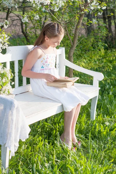 Leende flicka med öppen bok sitter på träbänk i vårträdgård — Stockfoto