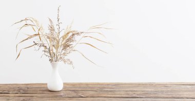 Beyaz arka plandaki ahşap masada vazoda kurumuş çiçekler.