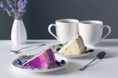 Böğürtlenli cheesecake ve arka plandaki gri duvardaki beyaz masada iki fincan çay.