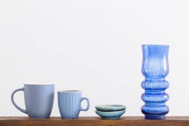 Beyaz zemin üzerinde ahşap rafta mavi bardaklar ve cam vazo