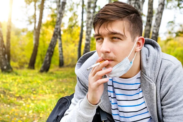 在秋天的公园里 戴着流感面具的年轻人在抽烟 — 图库照片
