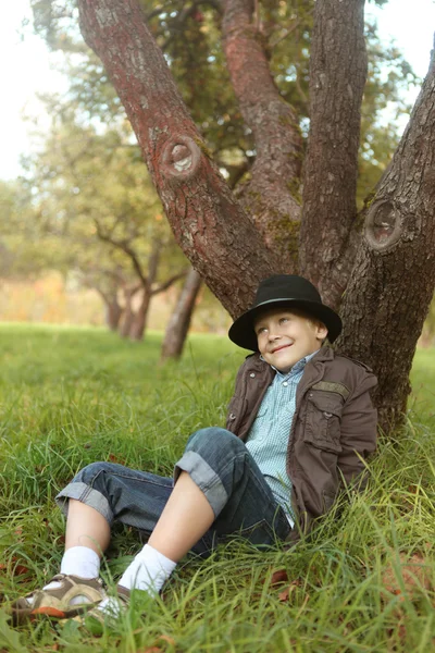 Lächelnder kleiner 6-jähriger Junge sitzt unter einem Baum im Park — Stockfoto