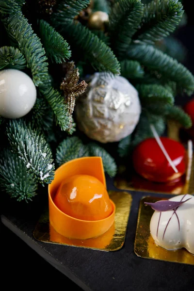 お菓子 様々な形や色のムースケーキ とチョコレートの作品とテーブル クリスマスのために装飾されたトウヒの枝は背景にあります ストック画像