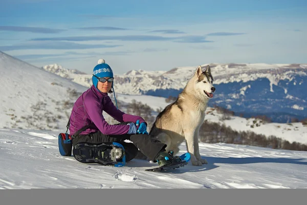 Una chica y un perro husky siberiano sentados en la nieve en las montañas —  Fotos de Stock