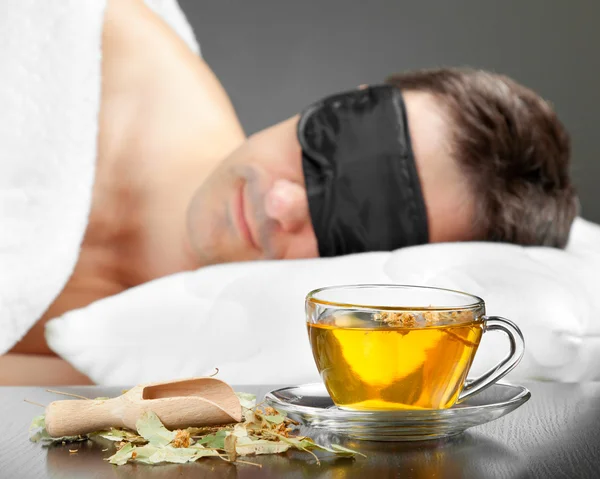 Man med sovande mask sömn på en säng, kopp örtte i den — Stockfoto