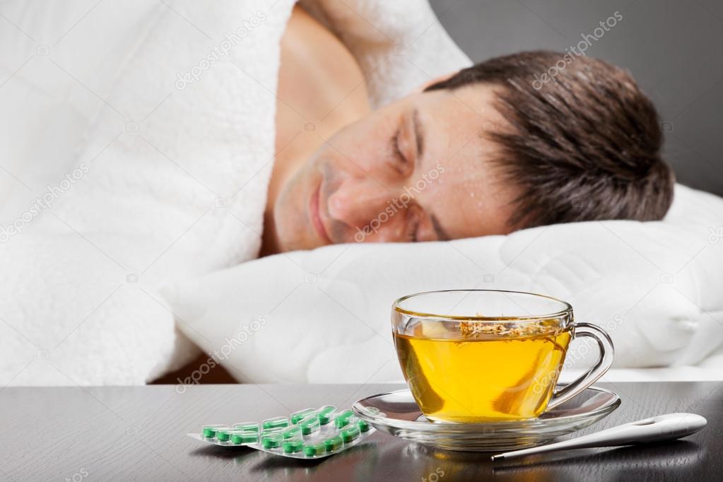 Sick man lying in bed with fever, cup of herbal tea, pills and