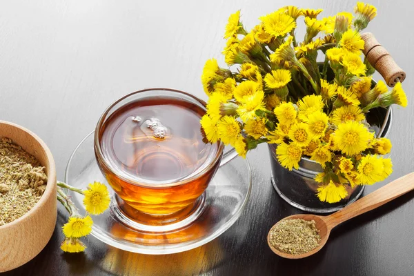 Healthy tea in glass cup closeup, bucket with coltsfoot flowers — Stock Photo, Image