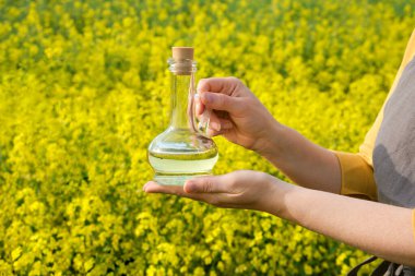 A woman stands in a rape field and holds a glass bottle or jug of rapeseed oil in his hands. Concept of canola oil production. clipart