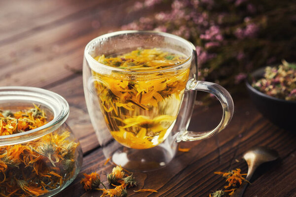 Glass cup of healthy calendula herbal tea, jar of dried calendula medicinal herbs and heather flowers on wooden table. Alternative herbal medicine.