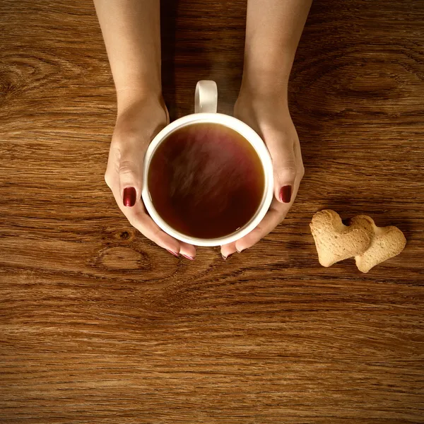 Mujer sosteniendo taza de té caliente con galletas en la mesa de madera —  Fotos de Stock