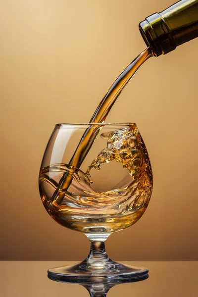Коньяк, що ллється з пляшки в склянку з бризкою на коричневому фоні — стокове фото