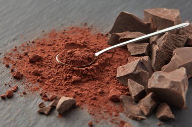 Kuru kakao tozu ve gri yüzeydeki organik çikolata parçalarının üzerine serpiştirilmiş parlak çay kaşığı.