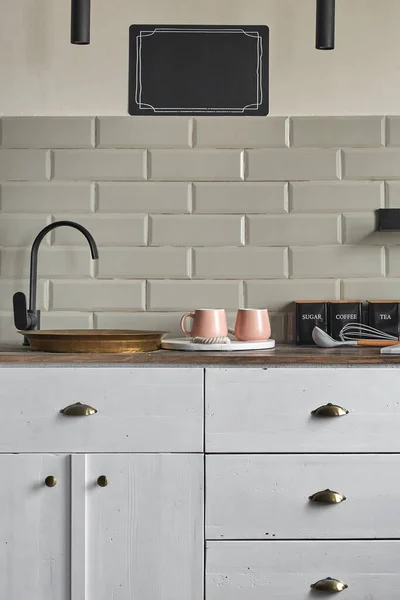 Old residential house kitchen cabinets with gray tiles wall, white cabinets, old copper sink in comfortable wooden counter pink mugs and tea and coffee containers. Country house kitchen concept.